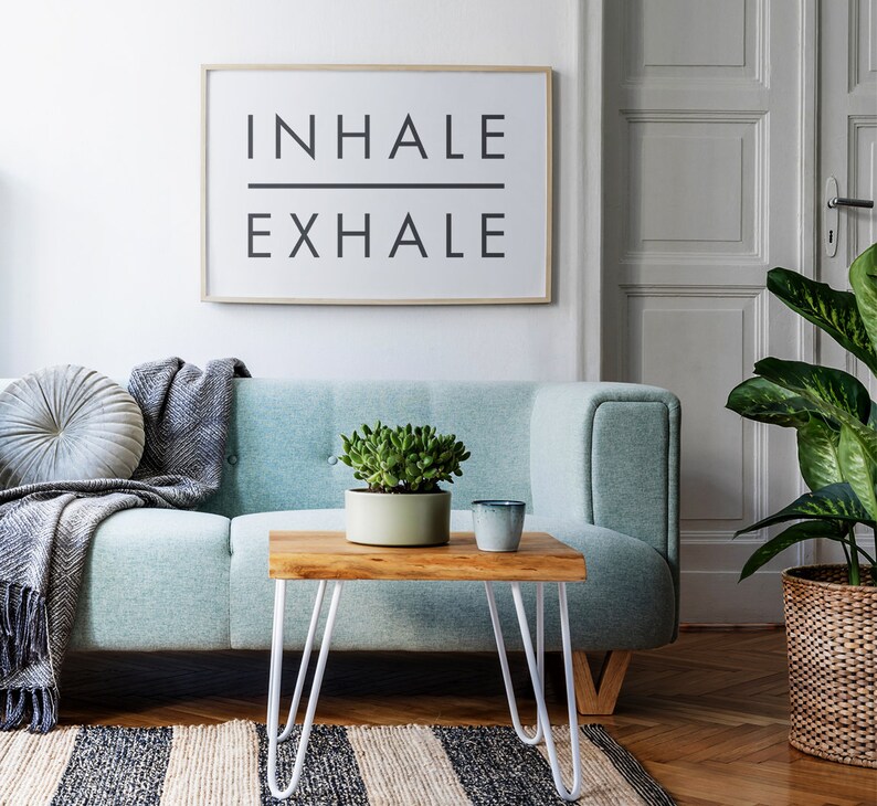 Op de afbeelding: Een lichtblauwe bank met een grijze deken en een rond grijs kussen. Een kleine houten tafel met witte haarspeldpoten staat voor de bank. Een potplant met een vetplant staat op de tafel, en een grijze keramische mok staat ernaast. Een ingelijst afdruk met de tekst "INHALE EXHALE" hangt aan de muur achter de bank.