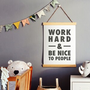 Puede incluir: Un p&oacute;ster enmarcado blanco con el texto "Work Hard & Be Nice To People" colgado en una pared gris con una guirnalda de papel de colores encima.