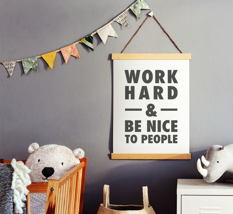 Puede incluir: Un p&oacute;ster enmarcado blanco con el texto "Work Hard & Be Nice To People" colgado en una pared gris con una guirnalda de papel de colores encima.