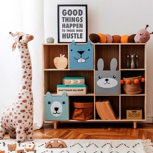May include: A wooden bookcase with various storage boxes and decorative items. The boxes feature animal faces. A stuffed giraffe and a sign that reads "GOOD THINGS HAPPEN TO THOSE WHO HUSTLE" are also present. A wooden car and blocks are on the floor.