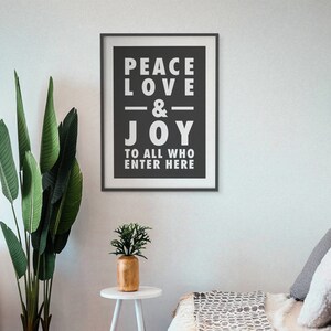 May include: Black and white framed print with the text "Peace Love & Joy to all who enter here". The print is hanging on a white wall with a green plant and a small table with a potted plant in front of it.