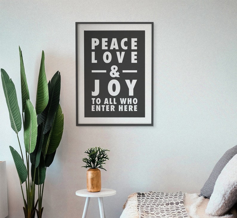 May include: Black and white framed print with the text "Peace Love & Joy to all who enter here". The print is hanging on a white wall with a green plant and a small table with a potted plant in front of it.