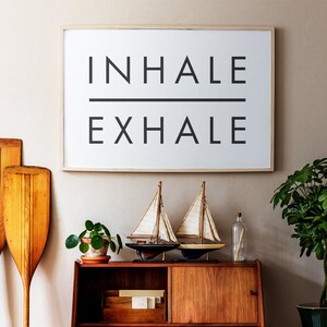 Op de afbeelding: Een ingelijst print met de woorden "INHALE EXHALE" in zwarte tekst op een witte achtergrond. De print hangt aan een beige muur boven een houten kast met twee zeilboten op de onderste plank.