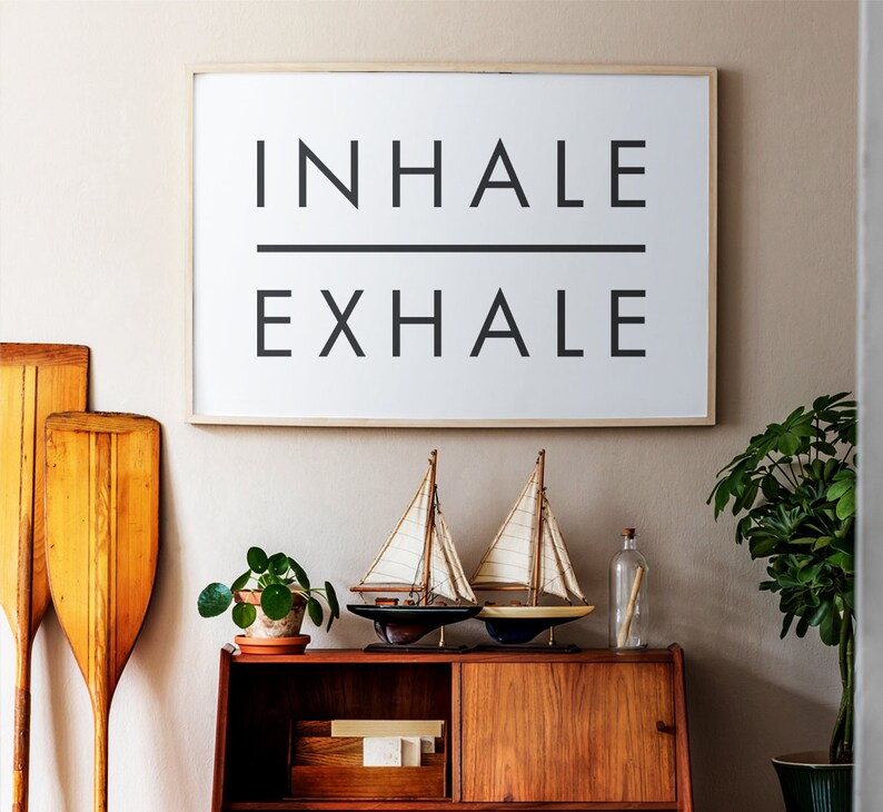 Op de afbeelding: Een ingelijst print met de woorden "INHALE EXHALE" in zwarte tekst op een witte achtergrond. De print hangt aan een beige muur boven een houten kast met twee zeilboten op de onderste plank.