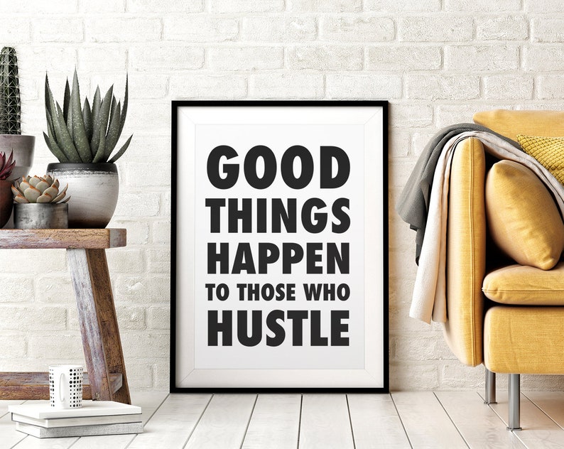 May include: A framed print with the text "GOOD THINGS HAPPEN TO THOSE WHO HUSTLE" in bold black letters. The print is in a black frame and is leaning against a white brick wall. A yellow sofa and plants are in the background.
