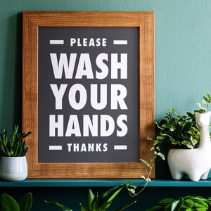 Puede incluir: Un letrero enmarcado con el texto "PLEASE WASH YOUR HANDS THANKS" en blanco sobre un fondo gris oscuro. El marco es de madera. El letrero est&aacute; en un estante con plantas.