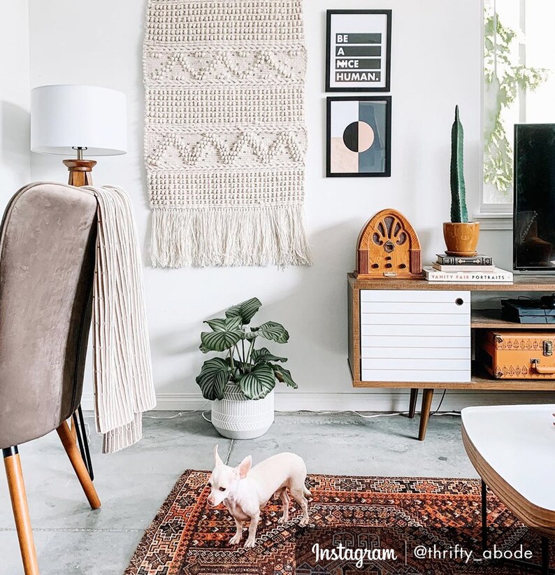 May include: A living room interior with a white wall, a white lamp, a brown leather chair, a white throw blanket, a white planter with a green plant, a white and brown mid-century modern cabinet, a vintage radio, a framed print with the text "Be a Nice Human", a framed print with a geometric design, a small white dog, and a brown and red patterned rug.