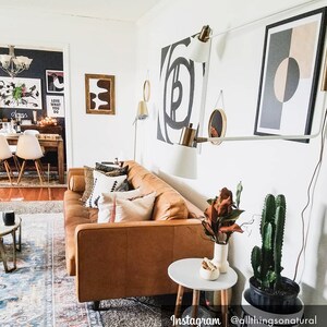 May include: A brown leather sofa with pillows in a living room with white walls. There are two black and white abstract paintings on the wall, a black and white geometric print, and a white lamp with a gold base. There is a small round table next to the sofa with a white vase and a cactus plant in a black pot.