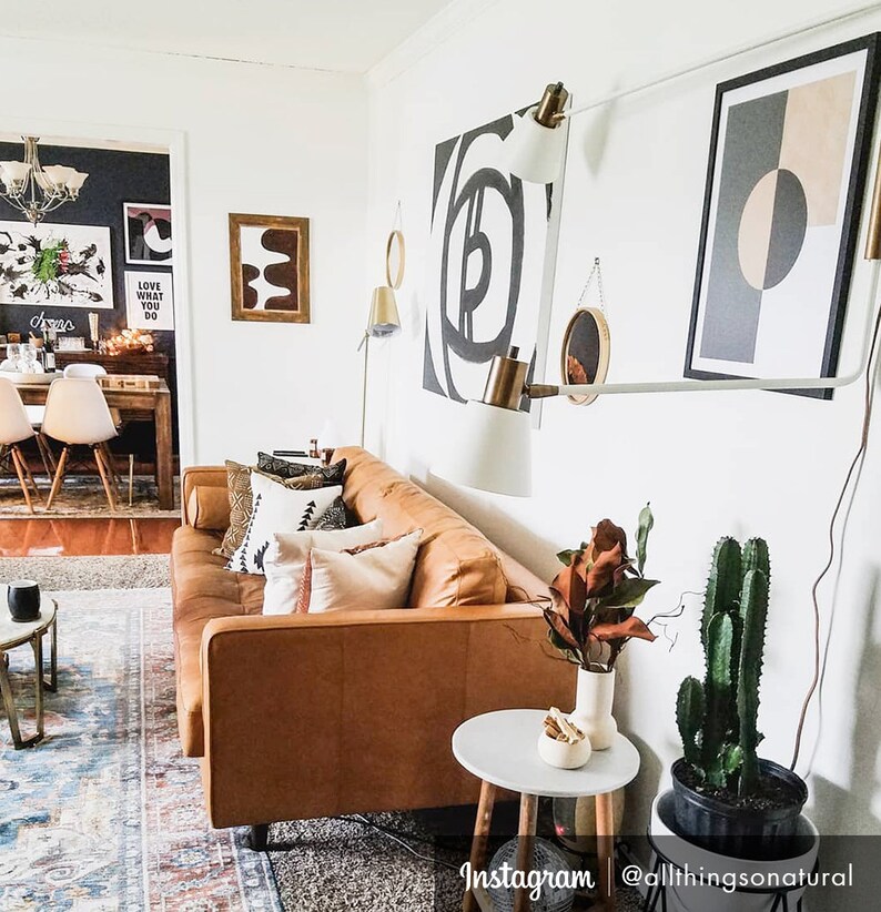 May include: A brown leather sofa with pillows in a living room with white walls. There are two black and white abstract paintings on the wall, a black and white geometric print, and a white lamp with a gold base. There is a small round table next to the sofa with a white vase and a cactus plant in a black pot.