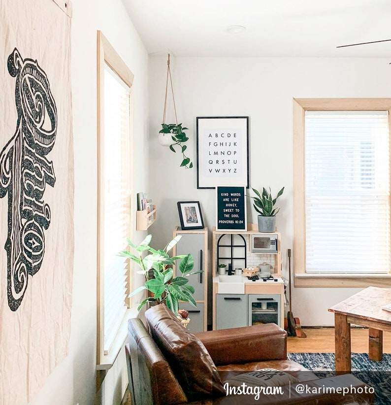 May include: A living room interior with a brown leather armchair, a play kitchen, and a white wall with black framed prints. The prints feature the alphabet and a quote about kind words.