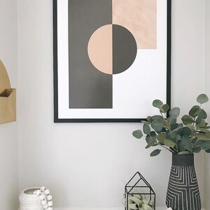 May include: A framed abstract print with geometric shapes in black, white, and pink. The print is hanging on a white wall above a white shelf with a white ceramic vase, a small potted plant in a black metal cage, and a vase with a black and white geometric pattern.