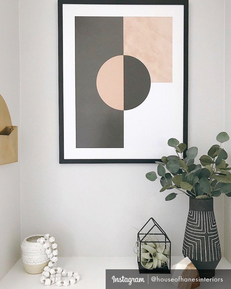 May include: A framed abstract print with geometric shapes in black, white, and pink. The print is hanging on a white wall above a white shelf with a white ceramic vase, a small potted plant in a black metal cage, and a vase with a black and white geometric pattern.