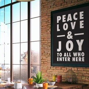 May include: A black framed sign with white text that reads "Peace Love & Joy To All Who Enter Here" hanging on a brick wall. The sign is in a modern loft style room with a large window and a wooden table with a vase of green plants.