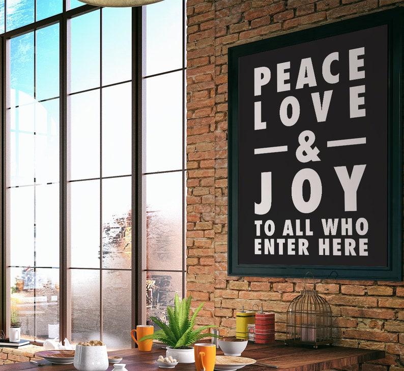 May include: A black framed sign with white text that reads "Peace Love & Joy To All Who Enter Here" hanging on a brick wall. The sign is in a modern loft style room with a large window and a wooden table with a vase of green plants.