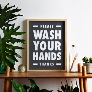 Puede incluir: Un letrero enmarcado con el texto "PLEASE WASH YOUR HANDS THANKS" en blanco sobre un fondo negro. El letrero se exhibe en una estanter&iacute;a de madera con plantas y una botella de vidrio.