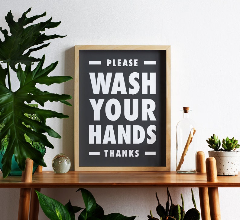 Puede incluir: Un letrero enmarcado con el texto "PLEASE WASH YOUR HANDS THANKS" en blanco sobre un fondo negro. El letrero se exhibe en una estanter&iacute;a de madera con plantas y una botella de vidrio.