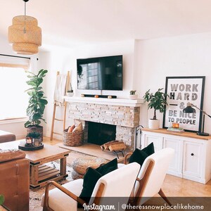 Puede incluir: Un interior de sala de estar con una chimenea de ladrillo blanco, un televisor de pantalla plana, un armario blanco con dos puertas y dos sillas blancas con cojines negros. La habitaci&oacute;n tiene un suelo de madera clara y una ventana grande con una persiana blanca. Un estampado enmarcado con el texto "Trabaja duro y s&eacute; amable con la gente" est&aacute; colgado en la pared.
