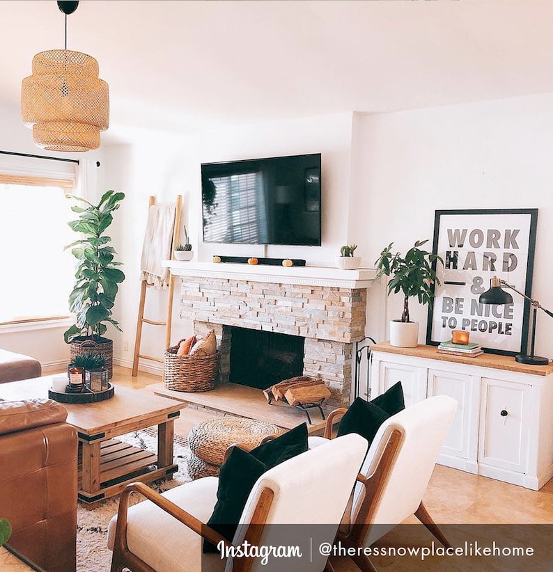 Puede incluir: Un interior de sala de estar con una chimenea de ladrillo blanco, un televisor de pantalla plana, un armario blanco con dos puertas y dos sillas blancas con cojines negros. La habitaci&oacute;n tiene un suelo de madera clara y una ventana grande con una persiana blanca. Un estampado enmarcado con el texto "Trabaja duro y s&eacute; amable con la gente" est&aacute; colgado en la pared.