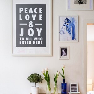 May include: A white framed print with the text "Peace Love & Joy to all who enter here" on a gray background. The print is hanging on a white wall with other framed prints and white tulips in vases.