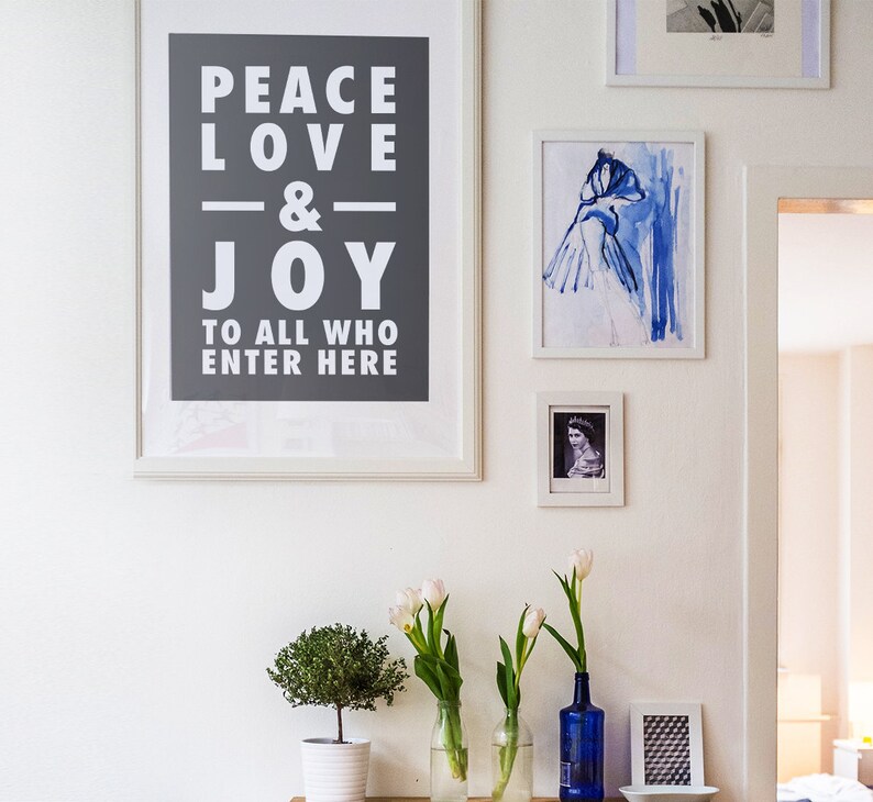 May include: A white framed print with the text "Peace Love & Joy to all who enter here" on a gray background. The print is hanging on a white wall with other framed prints and white tulips in vases.