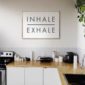 Op de afbeelding: Een witte keuken met een houten aanrechtblad en een ingelijst bord met de tekst "INHALE EXHALE".