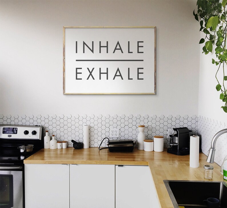 Op de afbeelding: Een witte keuken met een houten aanrechtblad en een ingelijst bord met de tekst "INHALE EXHALE".