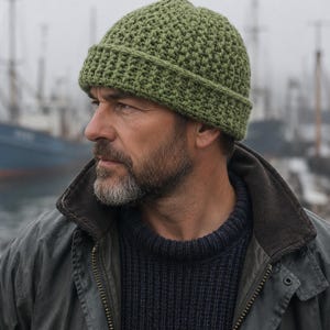 May include: A sage green beanie hat featuring a thick, textured crochet stitch pattern and a wide, folded brim. The hat is constructed from yarn with a visible, repeating knit-like texture, providing a dense and structured appearance.