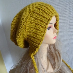 Könnte beinhalten: Eine senfgelbe Strickmütze mit einem langen, fließenden Schwanz. Die Mütze ist aus weichem, dickem Garn gefertigt und hat eine lockere Passform.