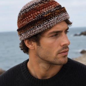 May include: A crocheted beanie featuring horizontal stripes in shades of brown, rust orange, cream, and dark charcoal. The hat has a thick, folded brim and a textured stitch pattern, crafted from yarn that blends these earthy tones throughout the design.