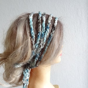 May include: A multi-strand braided headband crafted from variegated yarn in shades of blue, grey, and white. The accessory features several individual braided cords that wrap around the head, tied together at the side with loose ends.
