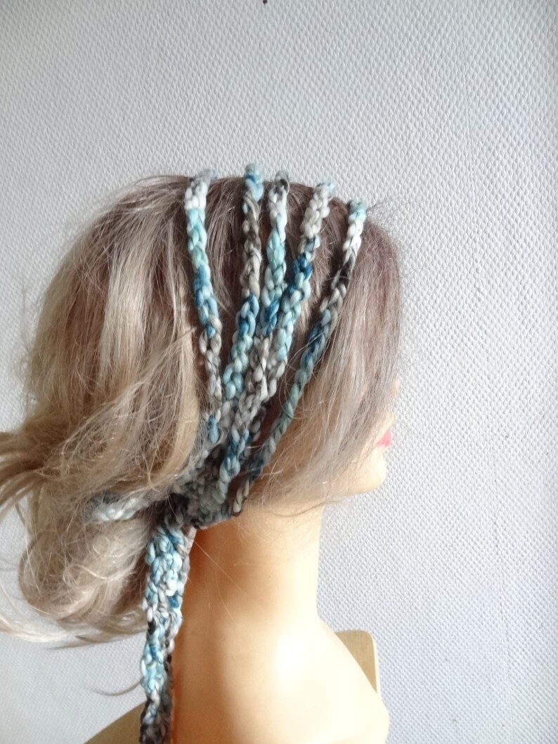 May include: A multi-strand braided headband crafted from variegated yarn in shades of blue, grey, and white. The accessory features several individual braided cords that wrap around the head, tied together at the side with loose ends.