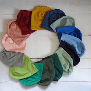 Op de afbeelding: Een cirkelvormige opstelling van vijftien zachte stoffen mutsen in een kleurenspectrum met mosterdgeel, donkerrood, lichtroze, grijs, limoengroen, smaragdgroen, bosgroen, saliegroen, marineblauw, hemelsblauw, koningsblauw en gedempt groenblauw.