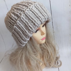 May include: A chunky knit beanie in a light silver-gray color. The hat features a prominent vertical ribbed texture throughout the fabric, with a thick, folded brim that sits securely around the crown. The yarn has a slight sheen to its finish.