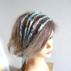 May include: A multi-strand braided headband featuring a variegated colour palette of light blue, white, and grey yarn. The accessory consists of several individual braided cords that wrap around the head, creating a layered and textured appearance.