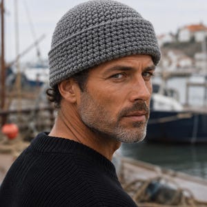 Puede incluir: Un gorro gris con un patrón de punto de ganchillo texturizado y un borde grueso doblado. El gorro está confeccionado con hilo denso y uniforme, con una corona redondeada y un ajuste ceñido que se sitúa justo por encima de las orejas.