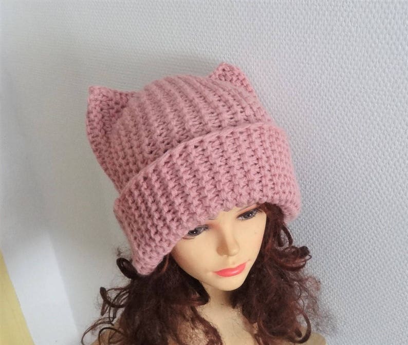 May include: A pink knitted beanie with cat ears. The beanie is made of a soft, textured yarn and has a simple, classic design.