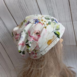 May include: A white beanie hat with a floral pattern featuring pink, purple, and yellow flowers with green leaves. The hat is made of a soft fabric and has a comfortable fit. The floral design adds a touch of elegance to the hat.
