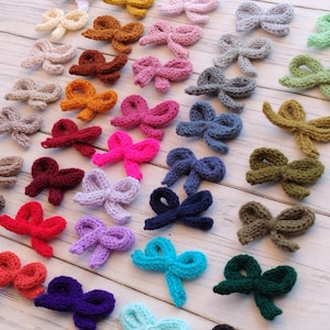 May include: A collection of small, knitted bows arranged in rows, featuring a wide variety of solid colours including white, brown, pink, grey, green, yellow, red, purple, blue, and black. Each bow is crafted with a textured, yarn-based knit stitch.