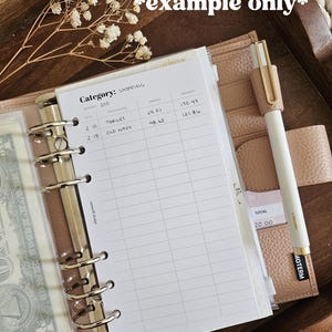 May include: A blush-colored planner with a pen and a shopping budget sheet. The sheet lists shopping expenses from Target and Old Navy. The pen is white with a gold clip and a black label that reads "MOTERM".