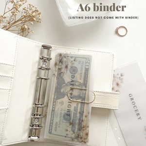 May include: White A6 binder with silver hardware, holding a clear plastic sleeve with floral accents and a twenty-dollar bill. The text "DATE NIGHT" is visible. The image also includes a ring and a "GROCERY" insert.