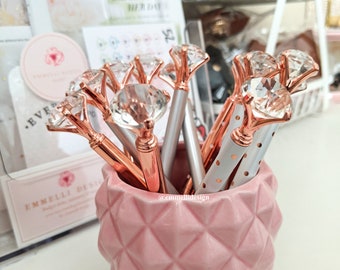 May include: A pink ceramic pineapple-shaped holder filled with rose gold pens topped with clear diamond-shaped accents. The pens are arranged in a cluster, with some of the diamond tops facing the camera. The text "Diamond Top Pen" is visible at the top of the image.