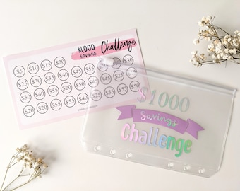 May include: A clear plastic zippered pouch with a pink and purple banner that reads "$1000 Savings Challenge". The pouch has six silver rings on the side. A pink and white sheet of paper with a grid of circles containing dollar amounts is visible in the background.
