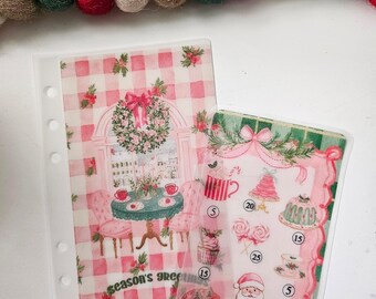 Könnte beinhalten: Zwei durchscheinende Planereinlagen mit Feiertagsgrafiken. Das linke Blatt zeigt einen rosa karierten Hintergrund, einen Kranz und ein Tischgedeck mit dem Text "season's greetings". Das rechte Blatt zeigt festliche Symbole, Zahlen und einen Teddybären mit dem Text "enjoy!".