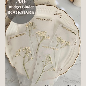 May include: A6 Budget Binder Bookmark with transparent pockets labelled "low priority", "sinking funds", "month ahead", and "high priority". Each pocket features dried white flowers. The text "Choose your Title" is at the bottom.