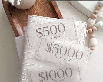 May include: Three vellum stickers with dollar amounts printed on them. The stickers are on a white leather notebook. The stickers say $500, $5000, and $1000.