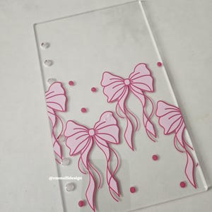 May include: Clear acrylic dashboard with pink bow and dot design. The text "Listing is for ONE Dashboard" is printed at the top. The design includes four bows and several small dots. The Etsy shop name "@emmelidesign" is printed at the bottom.