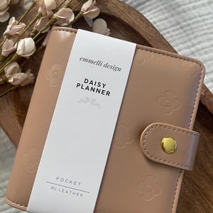 May include: A light brown PU leather pocket planner with embossed daisy patterns. The planner has a snap closure with a gold-colored button. The text "emmelii design" and "DAISY PLANNER" are printed on a white label. The planner is on a wooden tray.