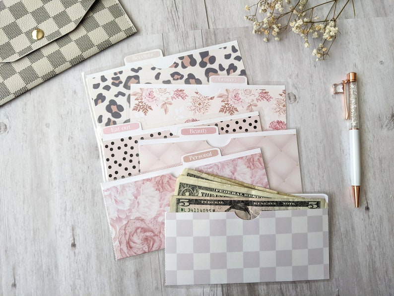 Puede incluir: Un juego de seis sobres de efectivo con estampado rosa y blanco con pesta&ntilde;as etiquetadas como "Recibos", "Comida", "Comer fuera", "Belleza", "Personal" y "Ahorros". Los sobres est&aacute;n abiertos y se pueden ver algunos billetes de d&oacute;lar en el sobre "Ahorros".