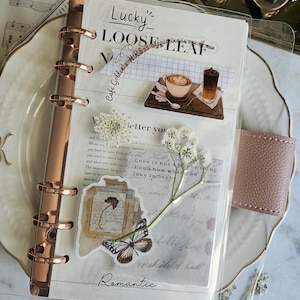 May include: A clear plastic planner dashboard featuring rose gold metal rings on the left side. The dashboard is decorated with stickers of coffee drinks, a butterfly, and pressed white flowers over printed text and vintage-style paper elements.