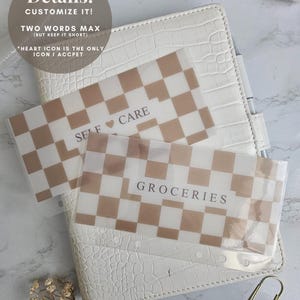 K&ouml;nnte beinhalten: Ein wei&szlig;er, strukturierter Planer mit zwei transparenten Eins&auml;tzen mit Muster. Die Eins&auml;tze haben ein braun-wei&szlig;es Schachbrettmuster und die W&ouml;rter "SELF CARE" und "GROCERIES". Eine goldene B&uuml;roklammer ist ebenfalls zu sehen.